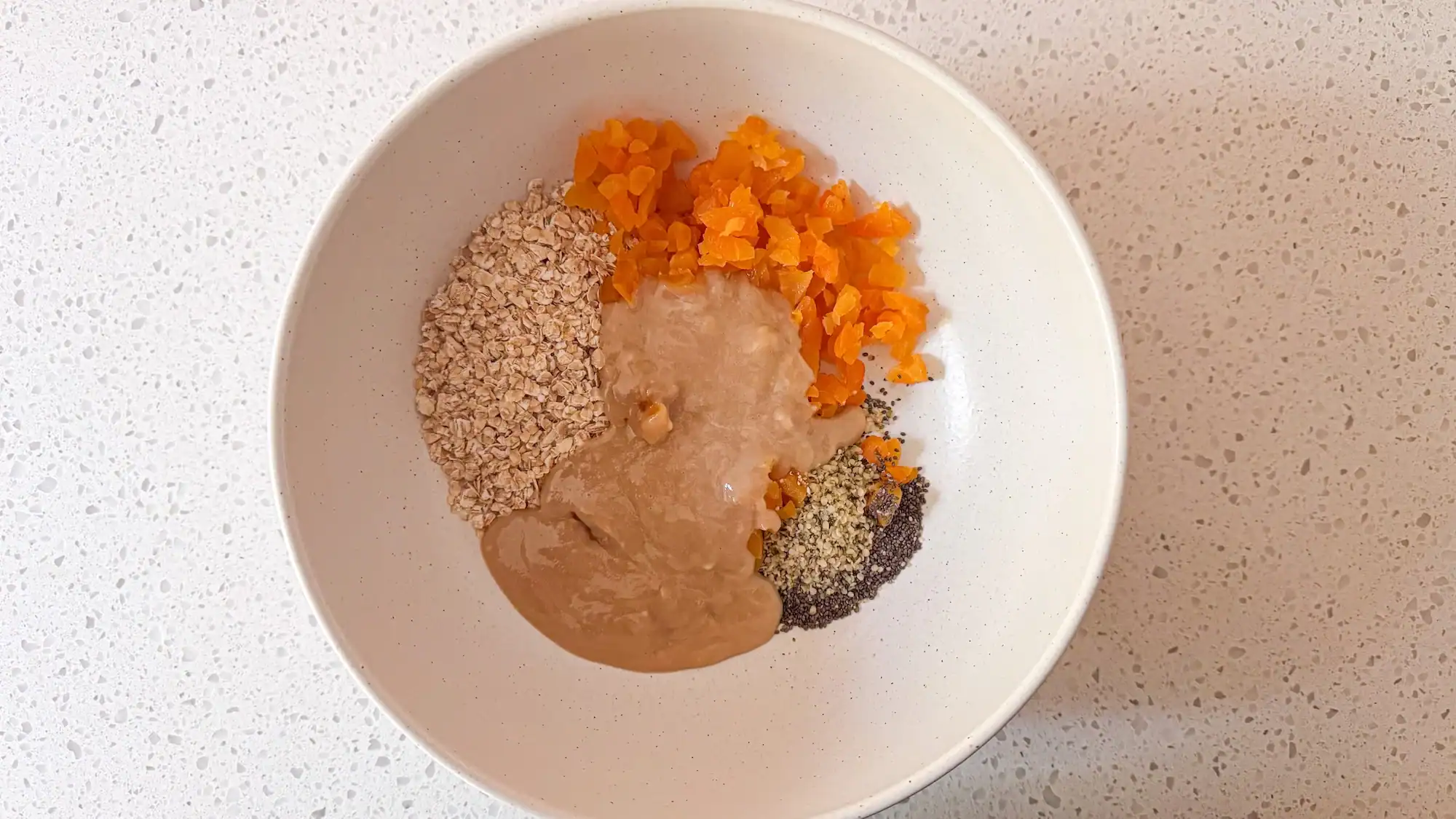 Step 3 for energy balls. Tahini mixture is added to the dry ingredients in a white bowl.