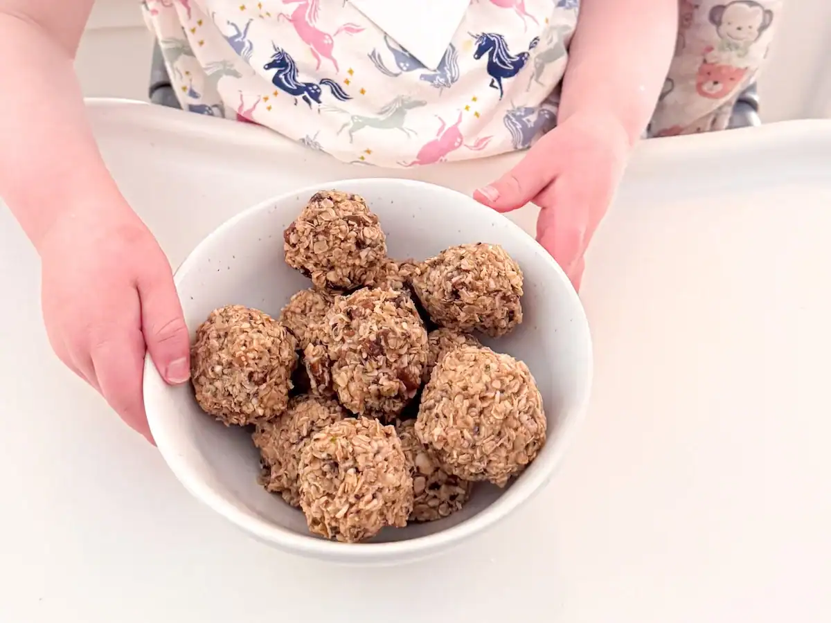Toddler at high chair holding bowl of energy balls.