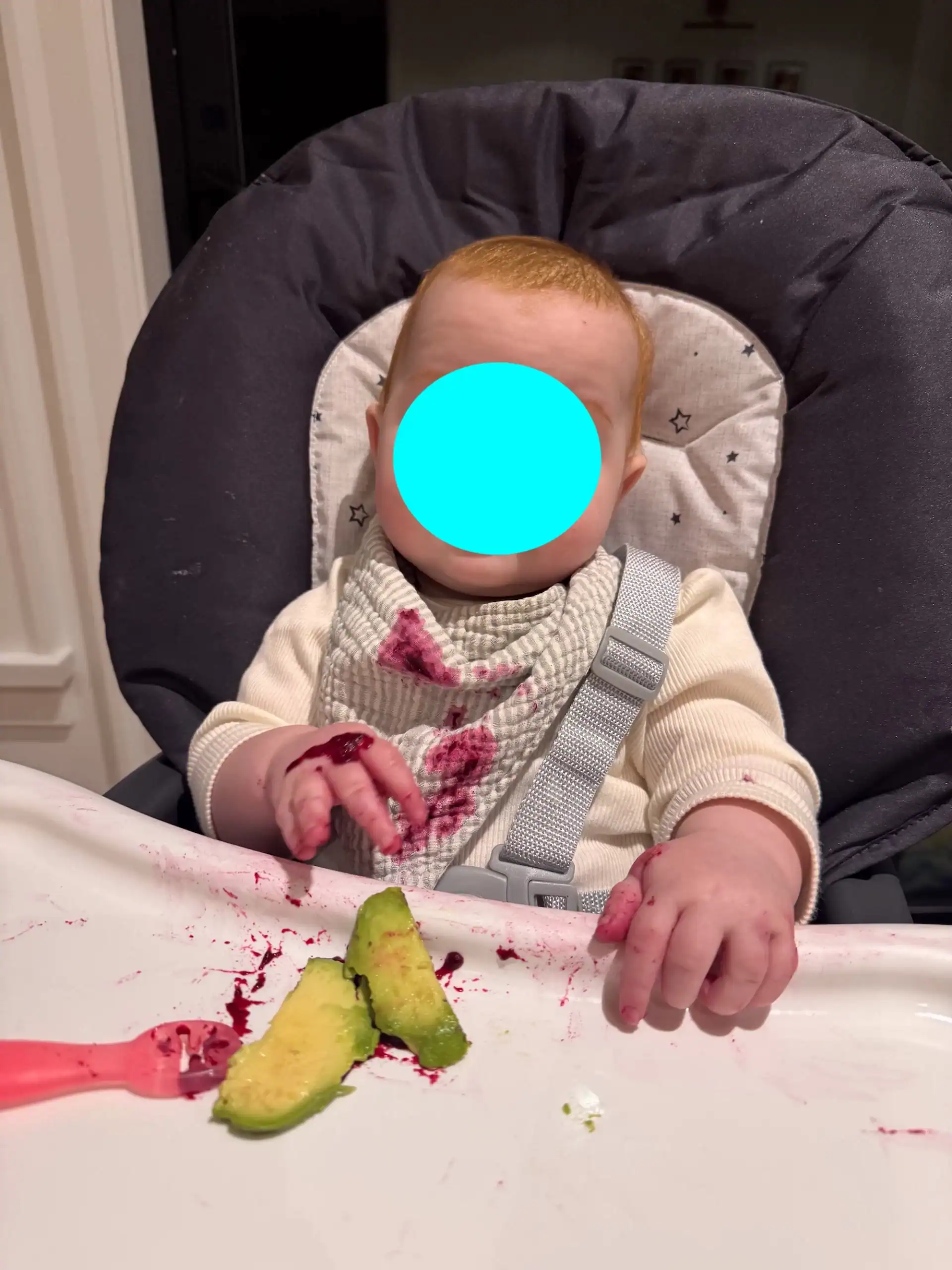 Baby in high chair eating avocado with blue circle covering face.