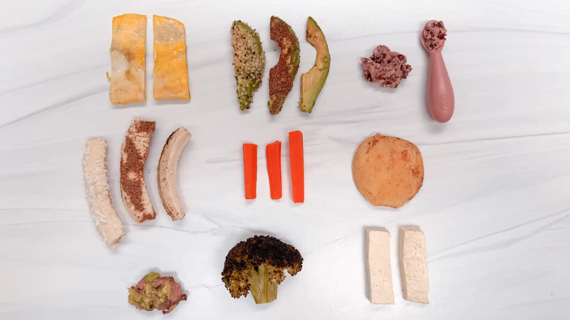 10 best first foods for baby on a white counter top. Includes carrots, apples, broccoli, omelette, tofu, chicken, avocado and beans.