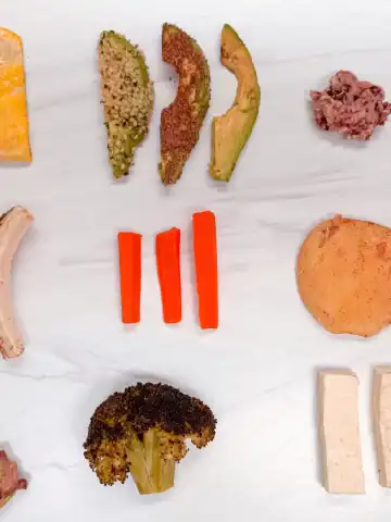 Top first foods for baby on a white counter. Includes foods like egg, avocado, carrots, apple, broccoli, tofu.